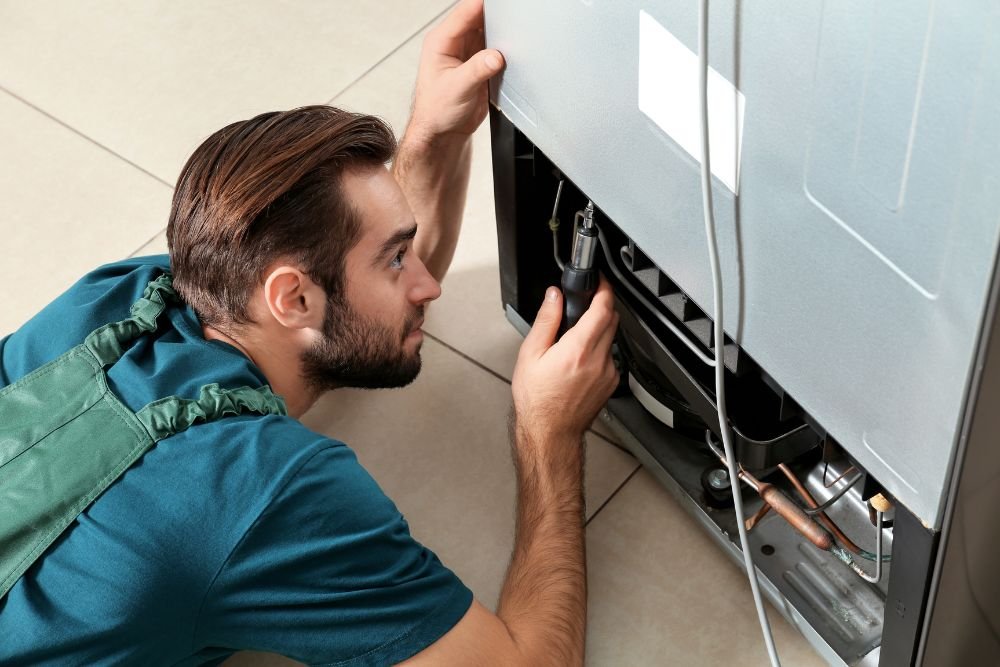 A man kneels on the floor, fixing a refrigerator, representing small appliance repair services in Everett, WA.